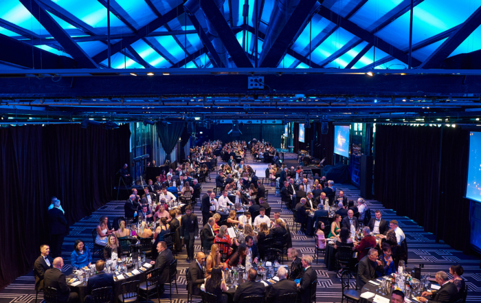 Large group of people sitting at tables in a large room. The room is decorated, it has tables with tablecloths and flowers. The people in the photo are all dressed up, and they are attending a formal event. Doltone House - Jones Bay Wharf is the Venue.